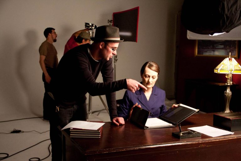 A man in a hat is pointing to a Laureen Smith at desk on set.
